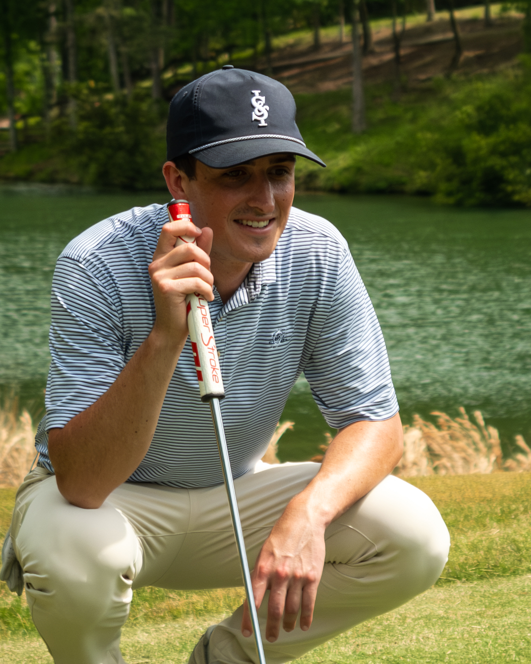 Golfer in a striped shirt and cap holding a golf club on a golf course with water and trees in the background.