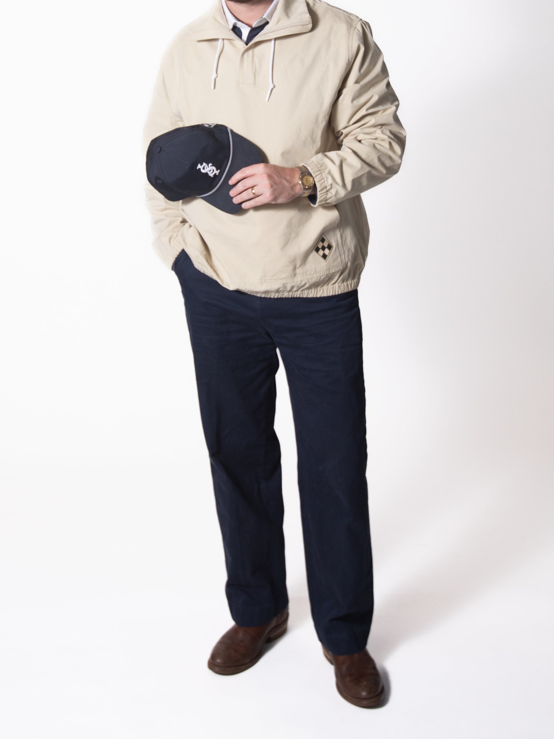 Person wearing a beige jacket and dark pants, holding a black cap with a white logo on a white background